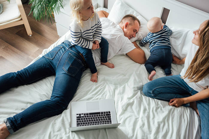 Happy young family sits together on bed and have fun time together at home. Tired dad trying rest while his mother and children playing