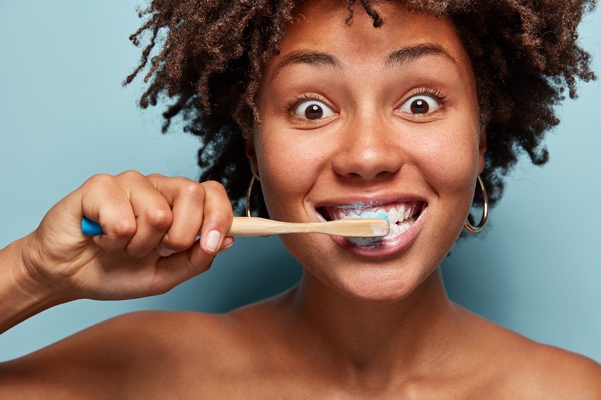 People and hygiene concept. Headshot of dark skinned woman with crisp hair, brushes and whitens teeth, cares for dental health, isolated over blue background, has morning routine. Stomatology concept