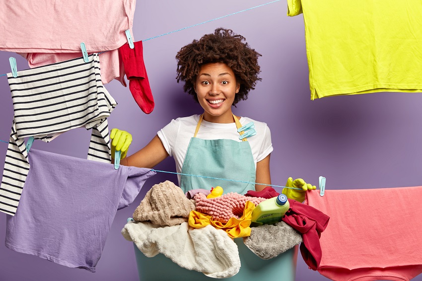 Smiling dark skinned housekeeper hangs clean clothes with clothespegs on washing line, happy to finish housework, laundry basket with cleaner near, wears domestic outfit, being busy at home.