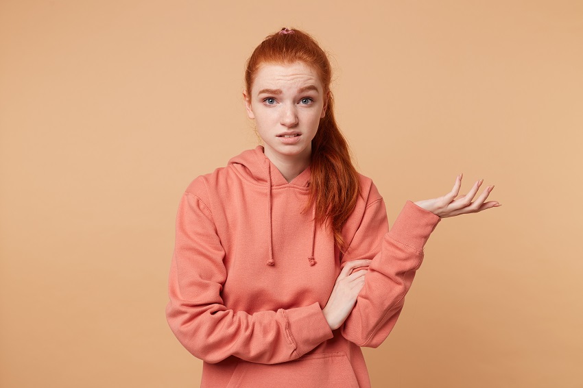 Dissatisfied red-haired girl with a tail dressed in hoodie with someone arguing one hand raised palm up trying to prove something, to defend their opinion, on beige studio background