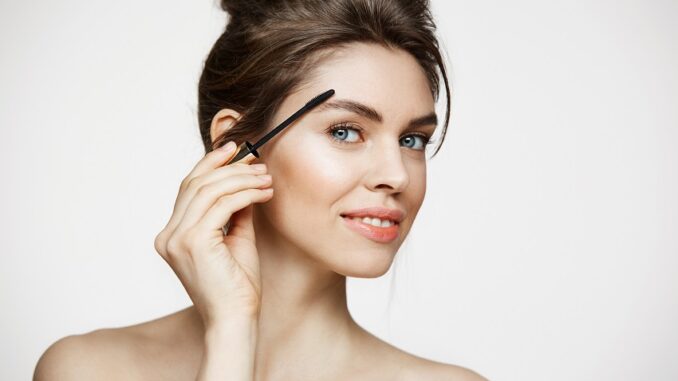 Young beautiful brunette girl smiling looking at camera painting eyebrows with mascara over white background. Beauty spa and cosmetology.